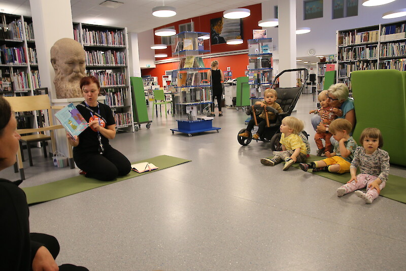 Päiväkotilaiset Jasper, Leevi, hänen takanaan oleva Nea, sylissä oleva Ellen, Oliver ja Amelia kuuntelevat mielenkiinnolla kirjastonhoitaja Anu Kokon lukemaa Muumi-satua.